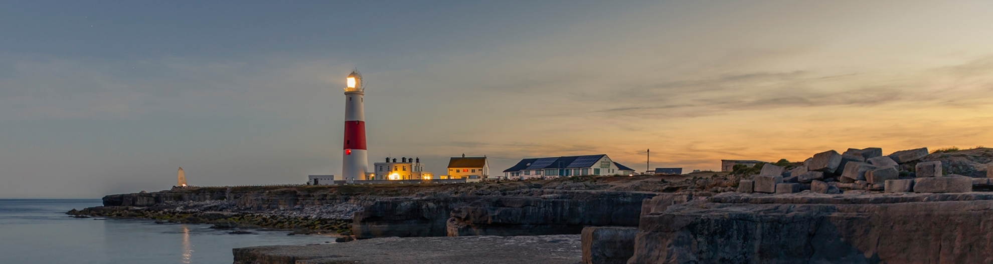 Illuminated Lighthouse By Sea Against Sky During Sunset